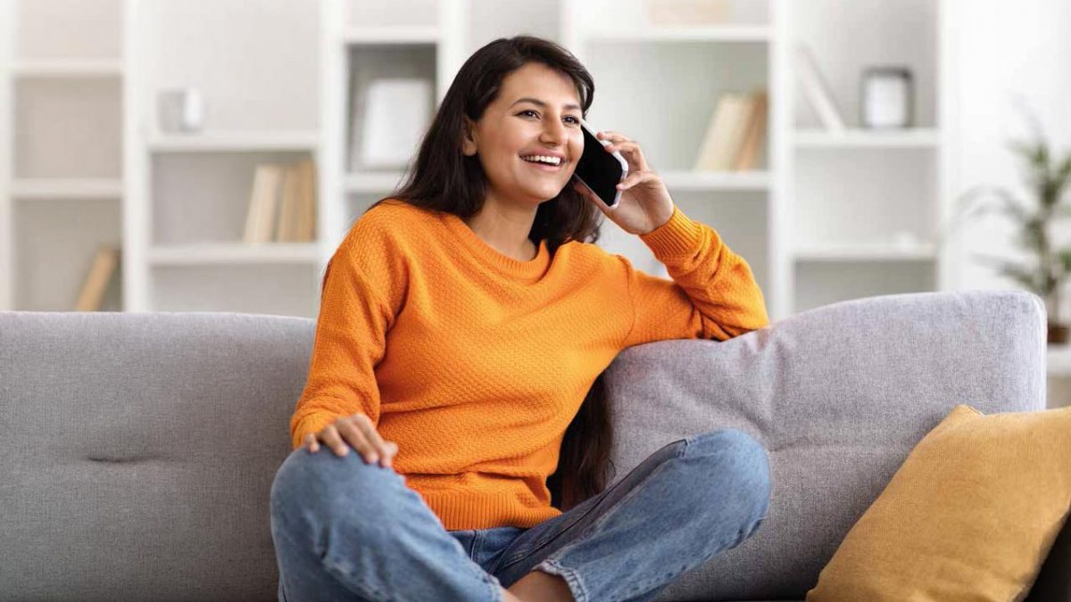 Lächelnde Person telefoniert entspannt auf einem Sofa in einem hellen Wohnraum.