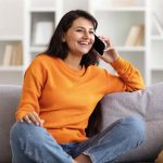 Lächelnde Person telefoniert entspannt auf einem Sofa in einem hellen Wohnraum.