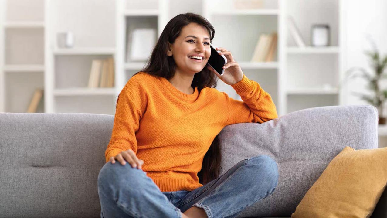 Lächelnde Person telefoniert entspannt auf einem Sofa in einem hellen Wohnraum.